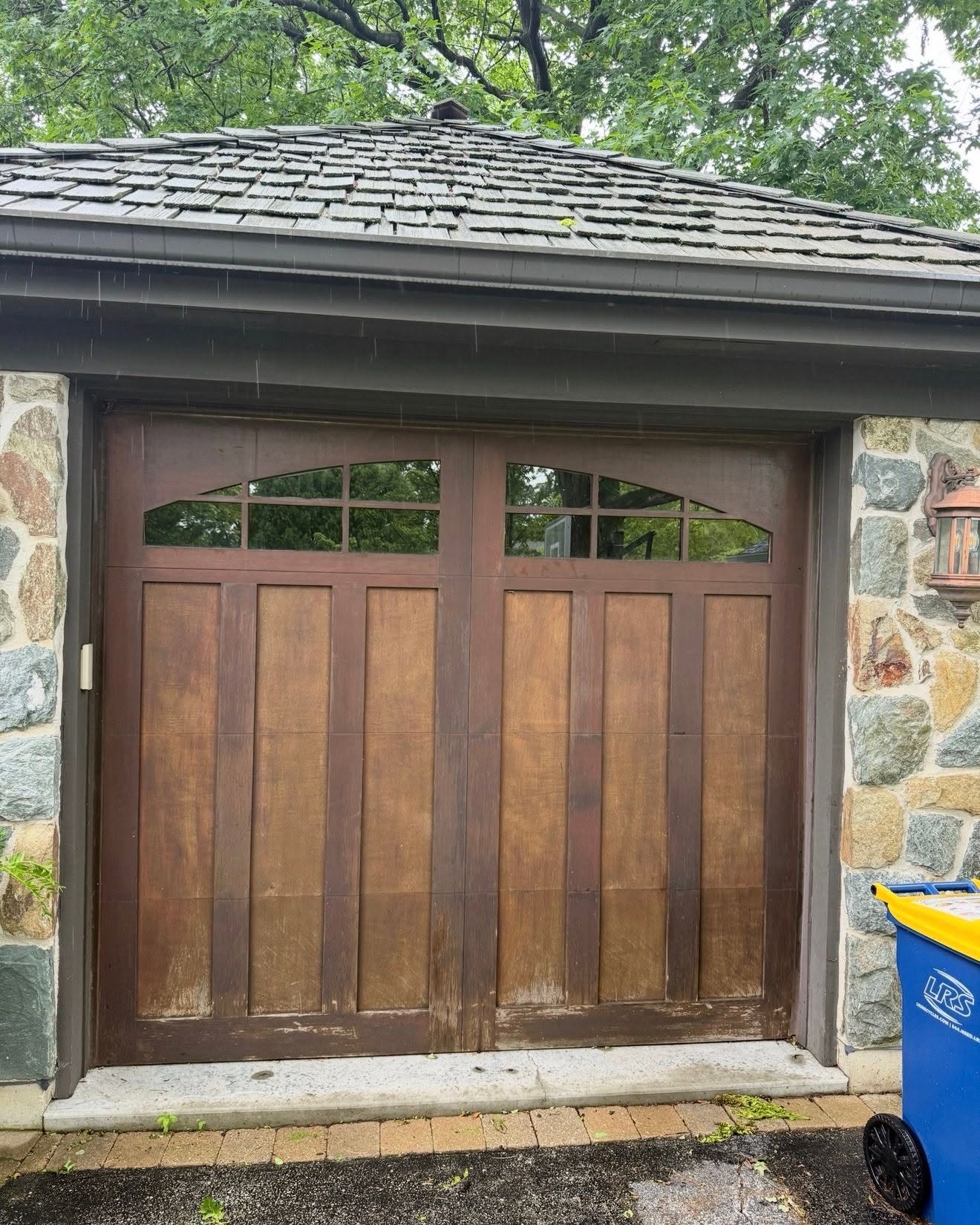 Before wood garage door staining in Glenview IL, faded and peeling stain with weathering at base panels on carriage-style door with stone surround