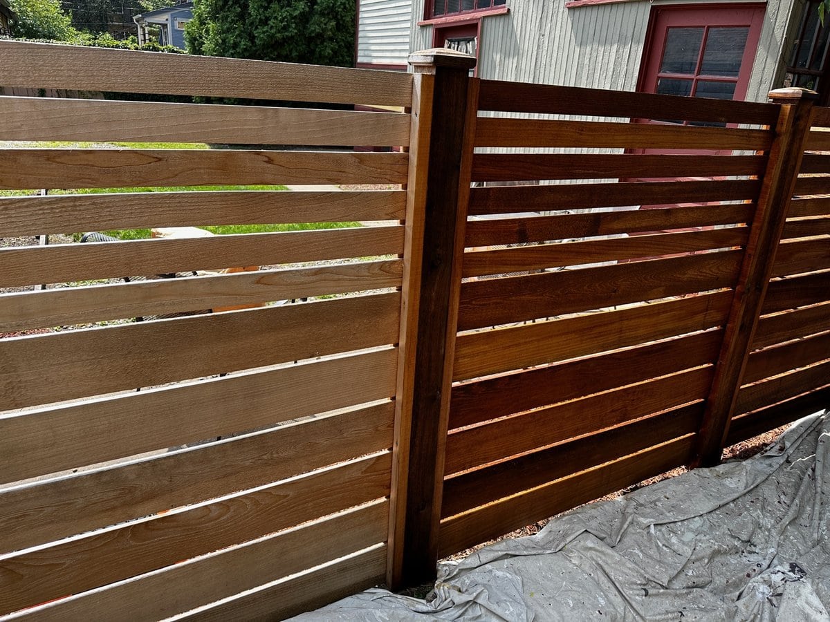 Plastic sheeting covering the ground along a horizontal wood fence during staining prep, Sunpainting project