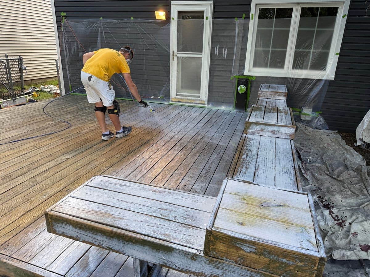 Sunpainting crew member in a yellow shirt and respirator spraying a weathered wood deck while the house wall behind is covered in plastic sheeting and tape
