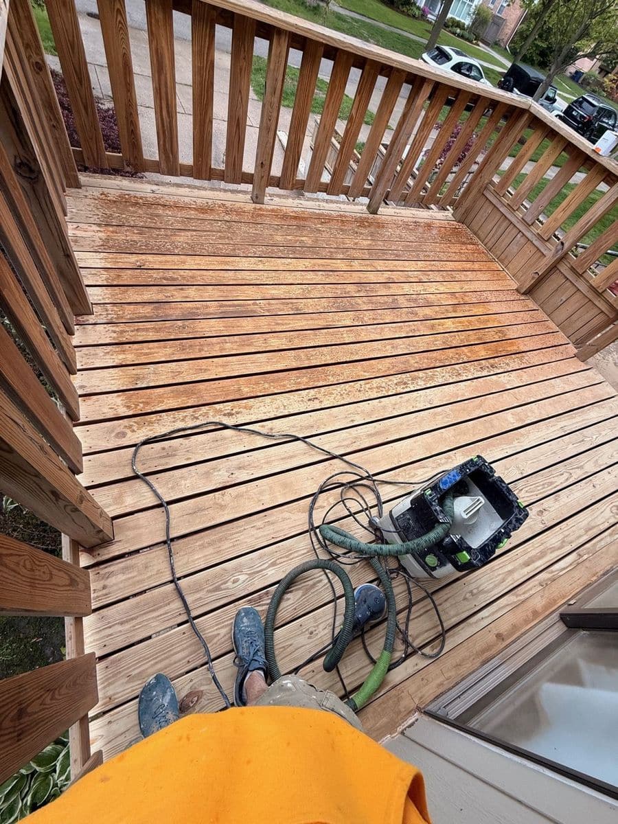 Wood deck boards being sanded with a sander connected to a vacuum hose, with a railing at the edge of the deck