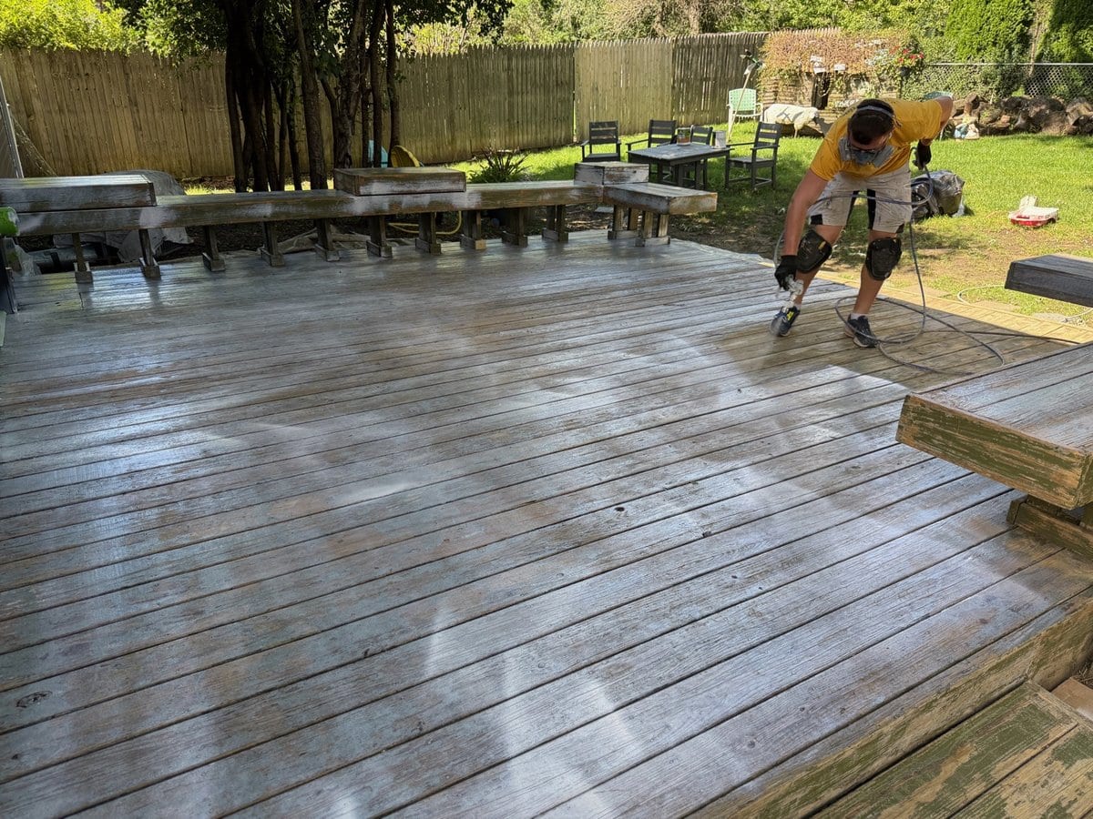 Sunpainting crew member in a yellow shirt spraying a large weathered wood deck with benches and a fenced yard behind