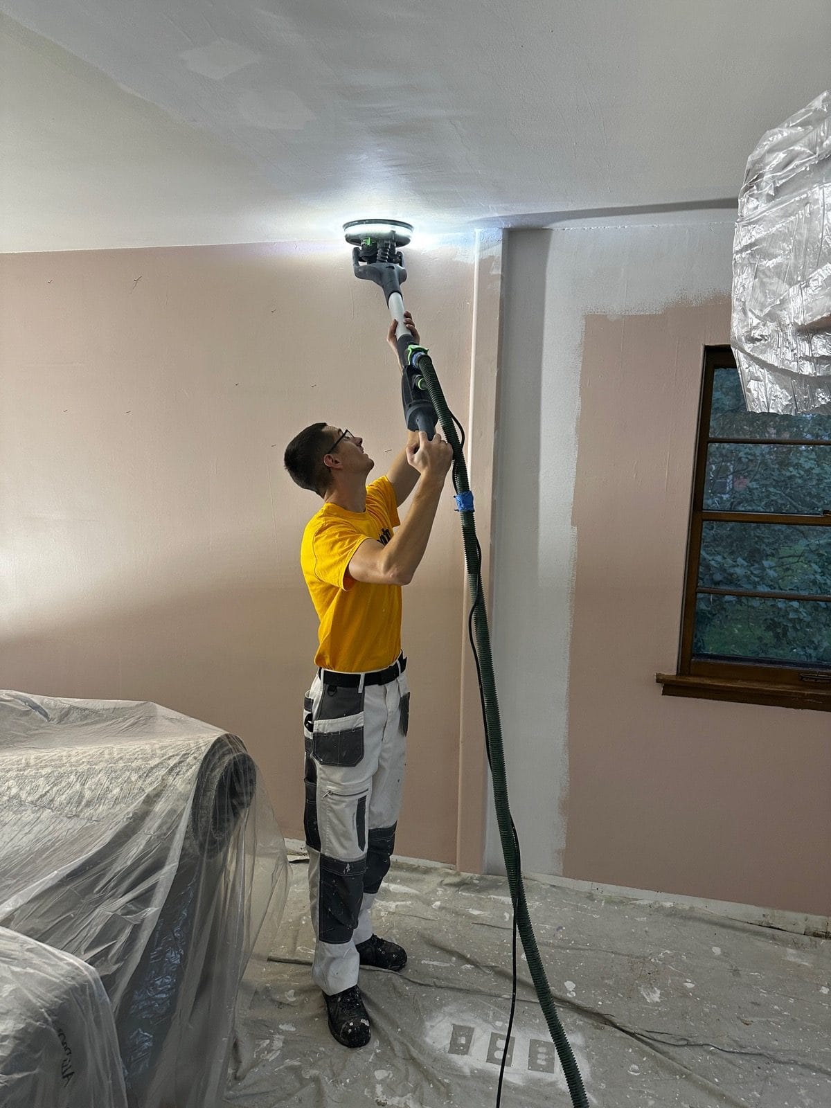 Sunpainting crew member sanding a ceiling with a dustless sander in a room with furniture covered in plastic and floors protected