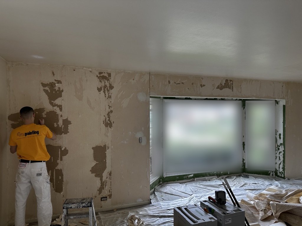 Sunpainting crew member stripping wallpaper from a living room wall, with plastic protecting the floor